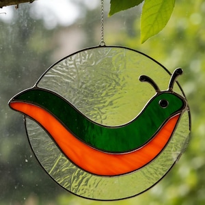 Op de afbeelding: Een glas-in-lood kunstwerk met een grillig slakkenontwerp. De slak heeft een groen lichaam met een oranje streep, tegen een achtergrond van getextureerd helder glas. Het stuk is cirkelvormig en hangt aan een ketting.