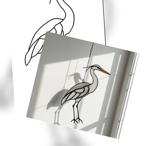 May include: Stained glass heron suncatcher with a white and grey body, black outline, and orange beak. The heron is suspended by a chain. A line drawing of the heron is in the background.