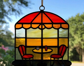Puede incluir: Atrapasoles de vidriera que representa una escena de café. El diseño presenta un toldo rojo y naranja, una mesa con dos sillas rojas, una taza y un vaso. Los colores incluyen rojo, naranja, amarillo y negro.