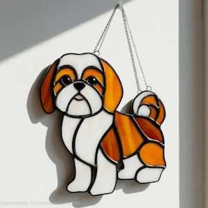 May include: A stained glass Shih Tzu dog suncatcher with a chain for hanging. The dog features white, orange, and brown glass pieces, with black outlines. A decorative piece for dog lovers. The dog's eyes, nose, and mouth are detailed.