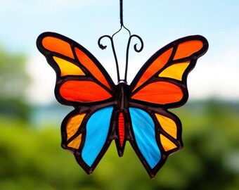 May include: A stained glass butterfly suncatcher with orange, yellow, blue, and black accents. The butterfly is hanging from a wire.