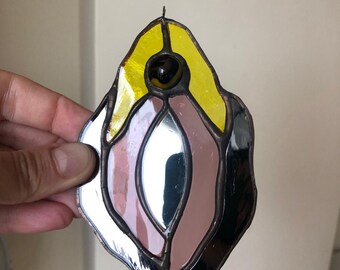 May include: Stained glass art piece with a unique design. The piece features yellow, brown, black, and mirrored glass sections, with a dark bead at the top. The edges are outlined with a dark metal. The piece is held by a hand.