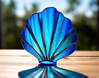 May include: A blue stained glass seashell with a suncatcher design. The shell is made of multiple pieces of glass that are joined together with lead. The shell is casting a shadow on a wooden surface.