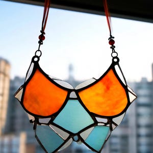 May include: A stained glass suncatcher in a geometric design, featuring orange, light blue, and white glass pieces. The suncatcher is suspended by a red ribbon and black metal chain, creating a decorative window hanging.