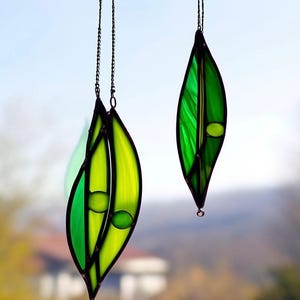 May include: Two stained glass leaf ornaments hanging from chains. The leaves are primarily shades of green and yellow, with dark outlines. Each leaf has a small, round, green accent. The background is blurred, suggesting an outdoor setting.