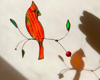 May include: A stained glass cardinal bird sculpture with a vibrant orange body, perched on a wire branch with green leaves and a red berry. The bird's shadow is cast on a white surface.