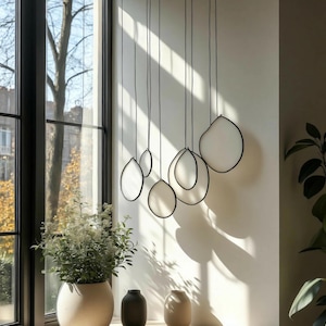 Könnte beinhalten: Eine moderne Leuchte mit mehreren weißen, oval geformten Schirmen, die von der Decke abgehängt sind. Jeder Schirm ist schwarz umrandet und hängt in unterschiedlichen Längen. Die Leuchte befindet sich vor einer weißen Wand, mit einem Fenster und Grün im Hintergrund.
