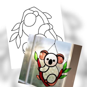 May include: Stained glass koala bear suncatcher. The koala has a white face, brown ears, and is perched on a red branch with green leaves. A black and white line drawing of a koala is in the background.