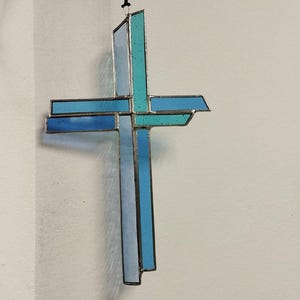 Puede incluir: Una cruz de vidrio coloreado con un esquema de color azul y turquesa. La cruz está colgada de una cuerda negra.
