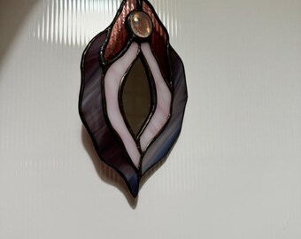 May include: Stained glass art piece in the shape of a stylized vulva. The piece features shades of purple, pink, and brown glass, with a clear, round gem at the top. The edges are outlined in black, and it hangs from a black string.