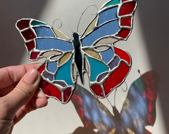 May include: A stained glass butterfly with a vibrant design. The wings feature sections of red, blue, teal, and yellow glass, outlined with silver solder. The butterfly is held up, casting a colorful shadow.