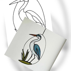 May include: A stained glass heron in an oval frame, with a black outline and a blue, white, and brown color scheme. The heron is perched on green reeds. A black and white line drawing of the heron is in the background.