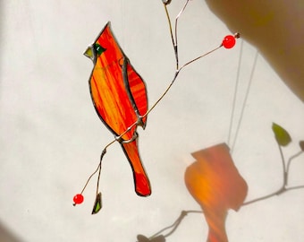 May include: A stained glass cardinal bird with red and orange hues perches on a wire branch with red bead berries and green leaves. The bird's shadow is cast on a white surface.