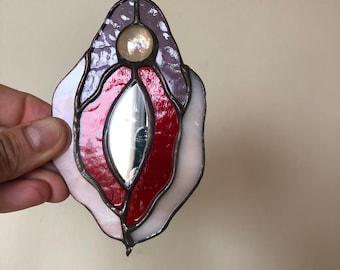 May include: A stained glass suncatcher in a stylized vulva shape. The piece features red, white, and purple glass with a central mirrored element. A small metal loop is at the top for hanging. The suncatcher is held by a hand.