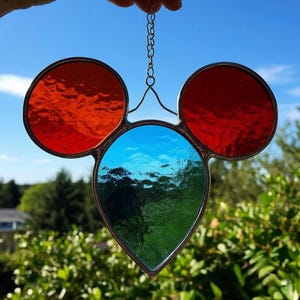 Op de afbeelding: Gebrandschilderd glazen suncatcher in een op Mickey Mouse geïnspireerd ontwerp. Voorzien van twee rode cirkels en een blauwe en groene druppelvorm, allemaal omlijst in donker metaal. Een kleine ketting is bevestigd om op te hangen.
