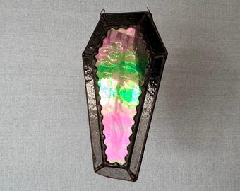 Puede incluir: Un adorno de vidrio negro en forma de ataúd con vidrio iridiscente rosa y verde en el interior.