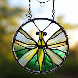 May include: A stained glass dragonfly suncatcher with a circular frame. The dragonfly features yellow, green, and white glass wings and body. The suncatcher is suspended by a black chain, perfect for adding a touch of nature-inspired art to any space.
