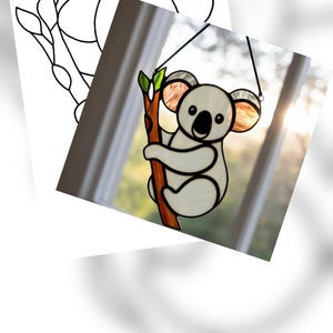 May include: Stained glass koala bear hanging decoration. The koala is white with black accents, hugging a brown branch with green leaves. The ears have a translucent orange and gray color. A black string is attached for hanging.