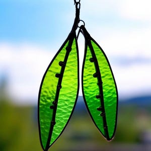 May include: Two leaf-shaped stained glass ornaments. The leaves are a vibrant green with a textured surface and black outlines. Small, dark beads are placed along a central vein. The background is blurred, with hints of blue and green.