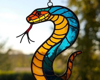 May include: A stained glass snake suncatcher in a curved pose, with a red tongue extended. The snake features a colour palette of blue, yellow, orange, and green, with black outlines. It hangs from a chain, perfect for window or wall decor.