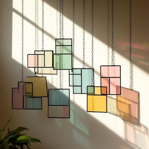 Puede incluir: Piezas de arte geométrico de vidrieras colgantes en varios colores pastel, incluyendo rosa, amarillo, verde y azul. Cada pieza está enmarcada en metal negro y suspendida por una cadena. La luz del sol proyecta sombras coloridas en la pared.