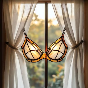 May include: A stained glass bra suncatcher with a warm color palette of amber, orange, and white. The decorative piece hangs in front of a window with white curtains. The bra design features geometric patterns and is suspended by a chain.