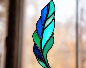 May include: Stained glass feather in shades of blue and green. The decorative piece is suspended by a small metal loop. The feather design features a variety of shapes and colors, creating a unique and artistic look.