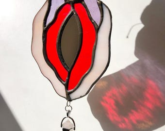 May include: A stained glass art piece in the shape of a vulva, featuring red, white, and lavender glass. A clear quartz crystal hangs from the bottom. The piece is held up by a hand, casting a shadow on a white surface.