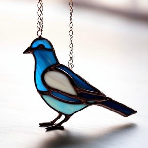 May include: A stained glass bird pendant with a blue and white design. The bird is hanging from a copper chain.