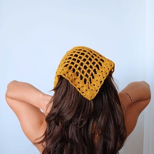 Puede incluir: Pañuelo de ganchillo amarillo mostaza con forma cuadrada. El cubrecabezas está hecho de un patrón similar a una red y se usa en la cabeza. El pañuelo es un accesorio de moda.