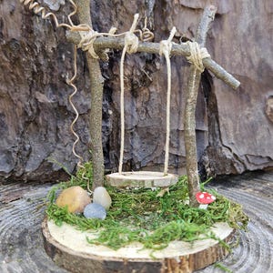 May include: A miniature fairy garden scene featuring a wooden swing suspended from branches. The swing sits on a bed of green moss, with small stones and a red and white mushroom. The base is a wooden slice.