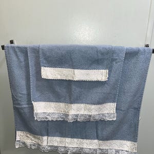 May include: A set of two dusty blue terry cloth towels hanging on a metal rack. Each towel features a wide band of white floral lace trim with a scalloped edge stitched horizontally across the lower section of the fabric.