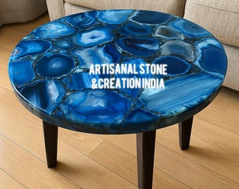 May include: Round blue agate stone coffee table with dark brown wooden legs. The tabletop features a mosaic of blue and white agate stones. The text "ARTISANAL STONE & CREATION INDIA" is visible on the table.