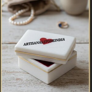 Puede incluir: Una caja de joyería rectangular blanca con ribetes dorados y un diseño de corazón rojo. La caja está abierta, revelando un interior de terciopelo rojo. El texto "ARTISANALSTONEINDIA" está impreso en la tapa. Un collar de perlas y un pequeño anillo están en el fondo.