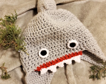 May include: Gray crocheted shark hat with red and white teeth and black eyes. The hat has a fin on top and a pointed hood.