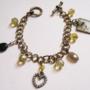 May include: A gold chain bracelet with multiple charms, including a heart, a square charm with a white image, and several clear and green beads.