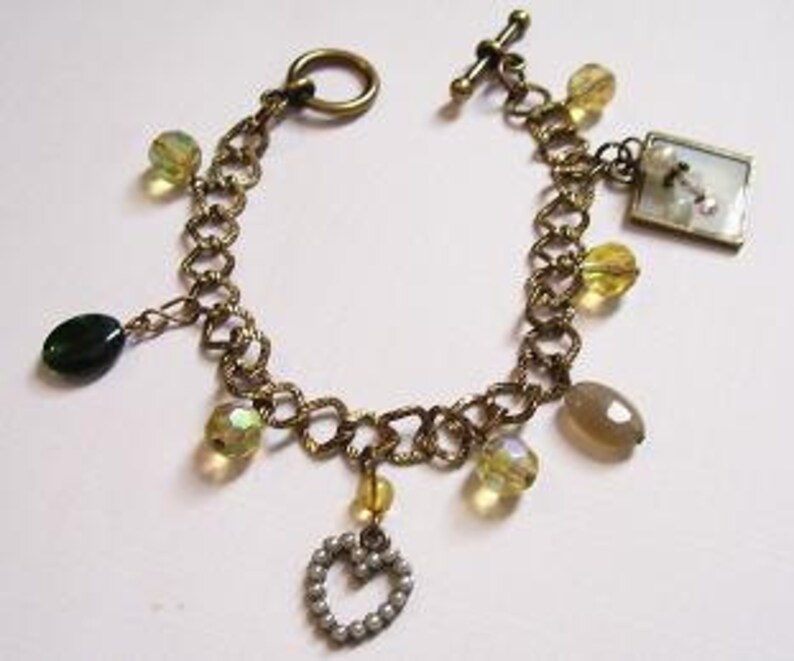 May include: A gold chain bracelet with multiple charms, including a heart, a square charm with a white image, and several clear and green beads.