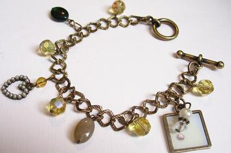 May include: A gold chain bracelet with a toggle clasp and multiple charms, including a small picture frame, a heart charm, and several clear and green glass beads.