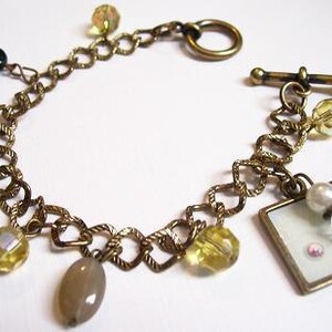 May include: A gold chain bracelet with a variety of colorful beads, including green, yellow, and white. The bracelet has a toggle clasp and a small charm with a white background.