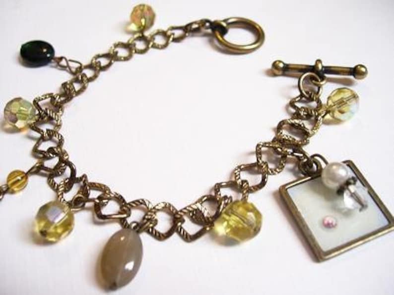May include: A gold chain bracelet with a variety of colorful beads, including green, yellow, and white. The bracelet has a toggle clasp and a small charm with a white background.