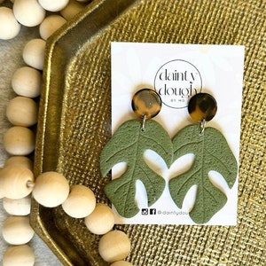 May include: A pair of green monstera leaf earrings with brown and gold accents. The earrings are on a gold tray with wooden beads. The text "dainty dough by mo" is on a white card behind the earrings.