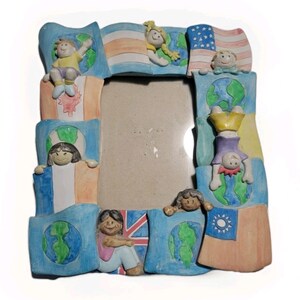 May include: A colorful picture frame with a central opening, decorated with flags and figures representing different countries. The frame features a blue border with various flags, including the American flag, and small, smiling figures. The frame is made of a ceramic material.