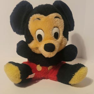 Puede incluir: Un peluche vintage de Mickey Mouse. El juguete presenta una cabeza y orejas negras, una cara amarilla y una nariz negra. Lleva pantalones cortos rojos con botones amarillos y pies amarillos. El juguete est&aacute; sentado con los brazos extendidos.