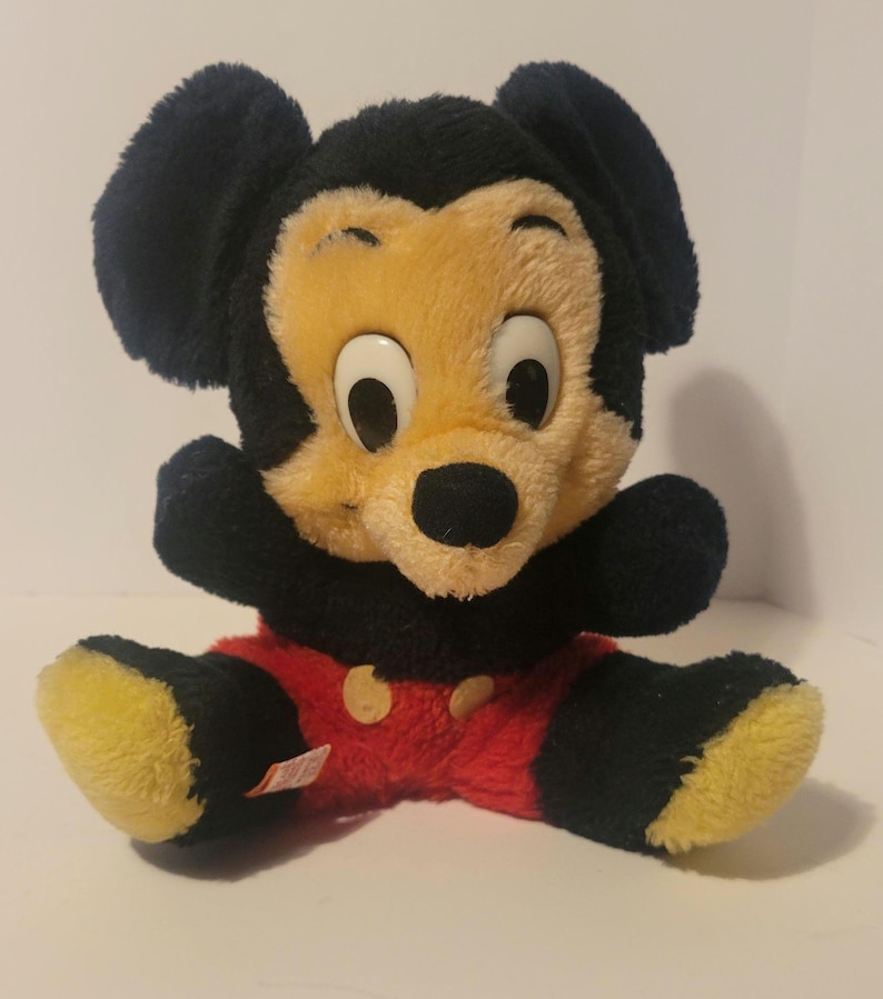 Puede incluir: Un peluche vintage de Mickey Mouse. El juguete presenta una cabeza y orejas negras, una cara amarilla y una nariz negra. Lleva pantalones cortos rojos con botones amarillos y pies amarillos. El juguete est&aacute; sentado con los brazos extendidos.