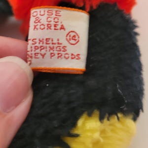 Puede incluir: Primer plano de un juguete de peluche con pelo rojo, negro y amarillo. Se ve una etiqueta con texto que incluye "HOUSE & CO. KOREA" y "SHELL LIPPINGS NEY PRODS". El dise&ntilde;o del juguete es parcialmente visible.