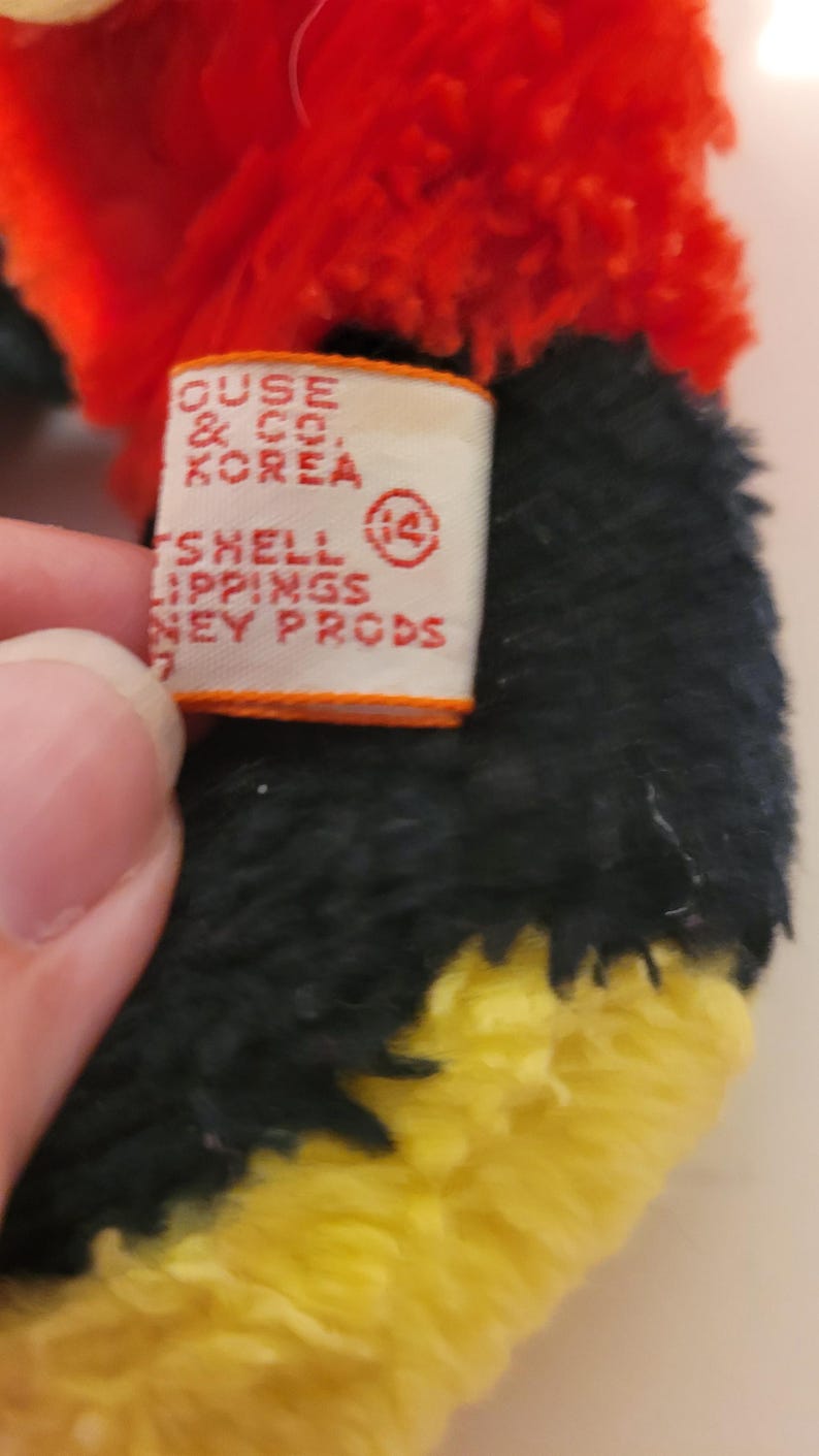 Puede incluir: Primer plano de un juguete de peluche con pelo rojo, negro y amarillo. Se ve una etiqueta con texto que incluye "HOUSE & CO. KOREA" y "SHELL LIPPINGS NEY PRODS". El dise&ntilde;o del juguete es parcialmente visible.