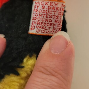 Puede incluir: Primer plano de un peluche con pelo negro, amarillo y rojo. Se ve una peque&ntilde;a etiqueta rectangular con el texto "MICKEY BY R. DAKIN PRODUCTS". El dise&ntilde;o del juguete es parcialmente visible, lo que sugiere un personaje de una popular serie de animaci&oacute;n.