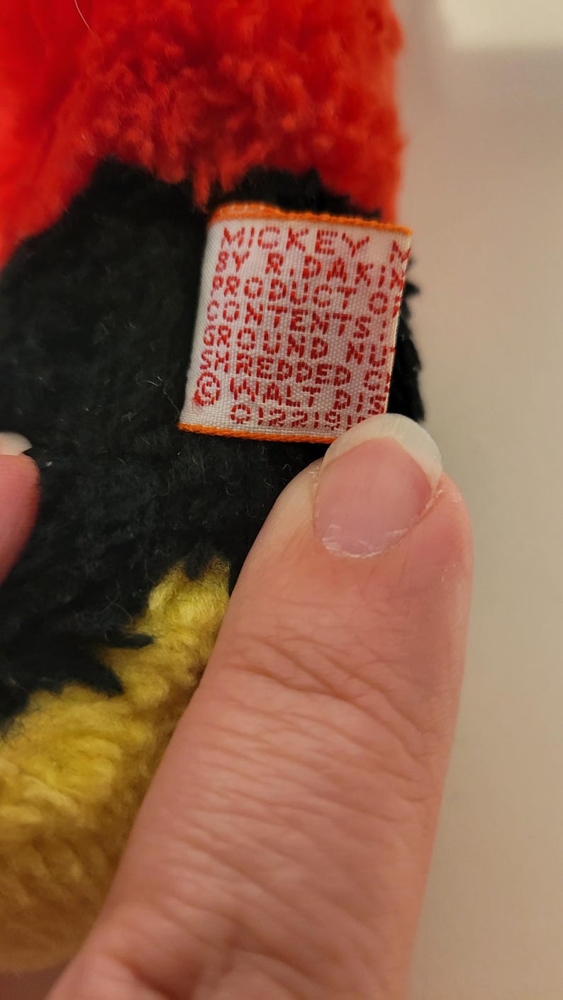 Puede incluir: Primer plano de un peluche con pelo negro, amarillo y rojo. Se ve una peque&ntilde;a etiqueta rectangular con el texto "MICKEY BY R. DAKIN PRODUCTS". El dise&ntilde;o del juguete es parcialmente visible, lo que sugiere un personaje de una popular serie de animaci&oacute;n.