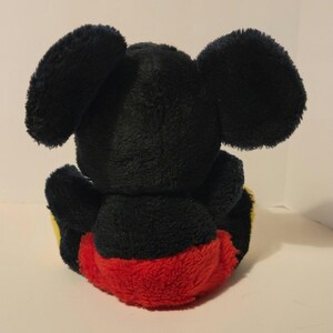 Puede incluir: Un peluche de un rat&oacute;n de dibujos animados, visto desde atr&aacute;s. El juguete tiene grandes orejas negras, un cuerpo negro y pantalones cortos rojos. Acentos amarillos son visibles en los pies. El juguete es suave y esponjoso.