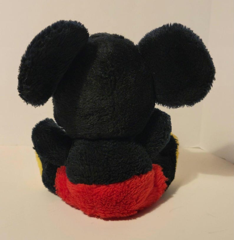 Puede incluir: Un peluche de un rat&oacute;n de dibujos animados, visto desde atr&aacute;s. El juguete tiene grandes orejas negras, un cuerpo negro y pantalones cortos rojos. Acentos amarillos son visibles en los pies. El juguete es suave y esponjoso.